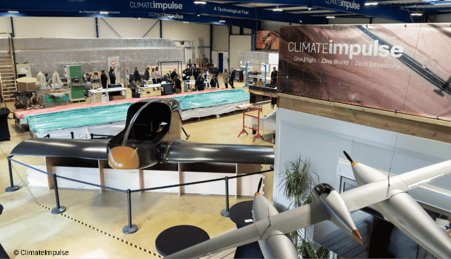 Climate Impulse Aircraft Hangar and Prototyping Workshop Large industrial aircraft hangar and workshop featuring prototypes of the Climate Impulse hydrogen airplane project. In the foreground, a sleek black fuselage mock-up and a twin-fuselage scale model are displayed behind stanchions, while team members gather in the background beneath large banner signs reading "CLIMATEimpulse – One Flight | One World | Zero Emissions."